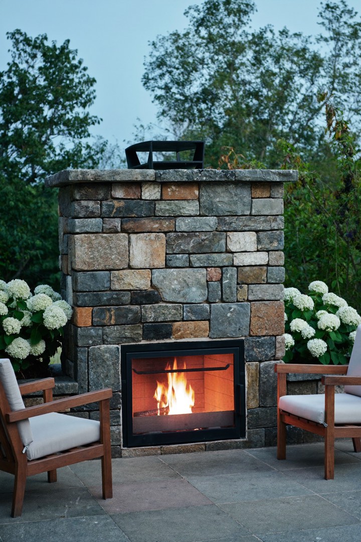 A fieldstone outdoor fireplace with a wood fire and two teak chairs at dusk.
