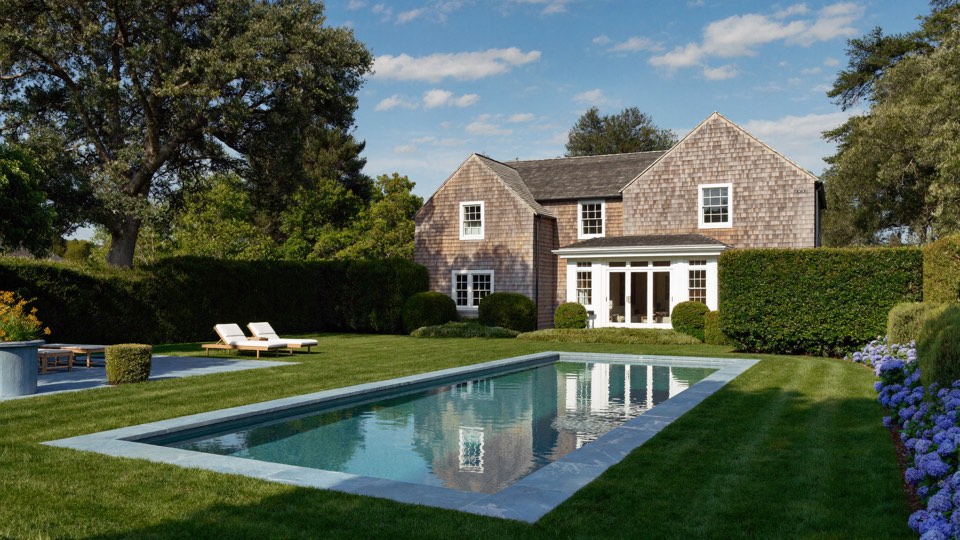 A long pool with pale stone coping reflecting a grey shingled house.