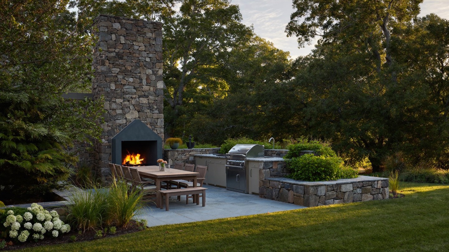 A stone terrace at golden hour with a lit fieldstone fireplace, a dining table and a built-in grill, under mature oaks.