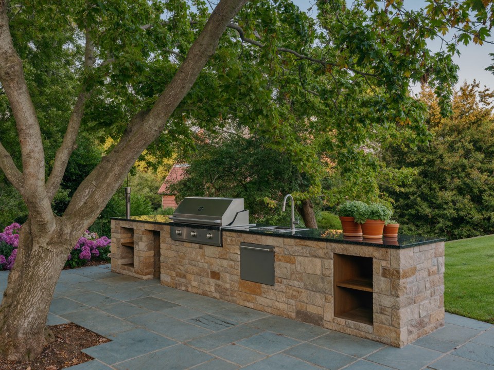 A built-in outdoor kitchen with a stone base, dark counter, grill and sink beneath a maple tree.