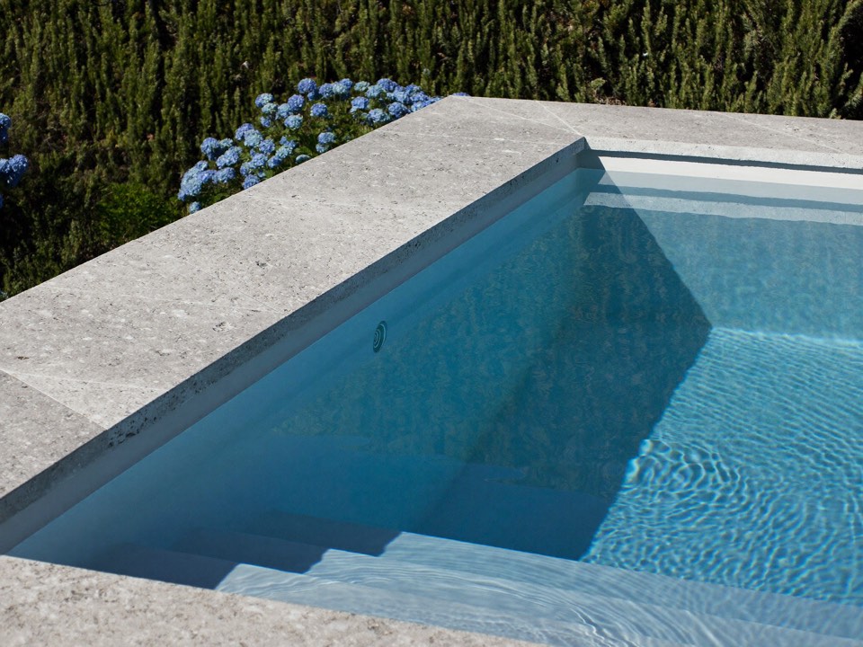 The corner of a pool with wide pale stone coping, clear blue water and hydrangeas beyond.