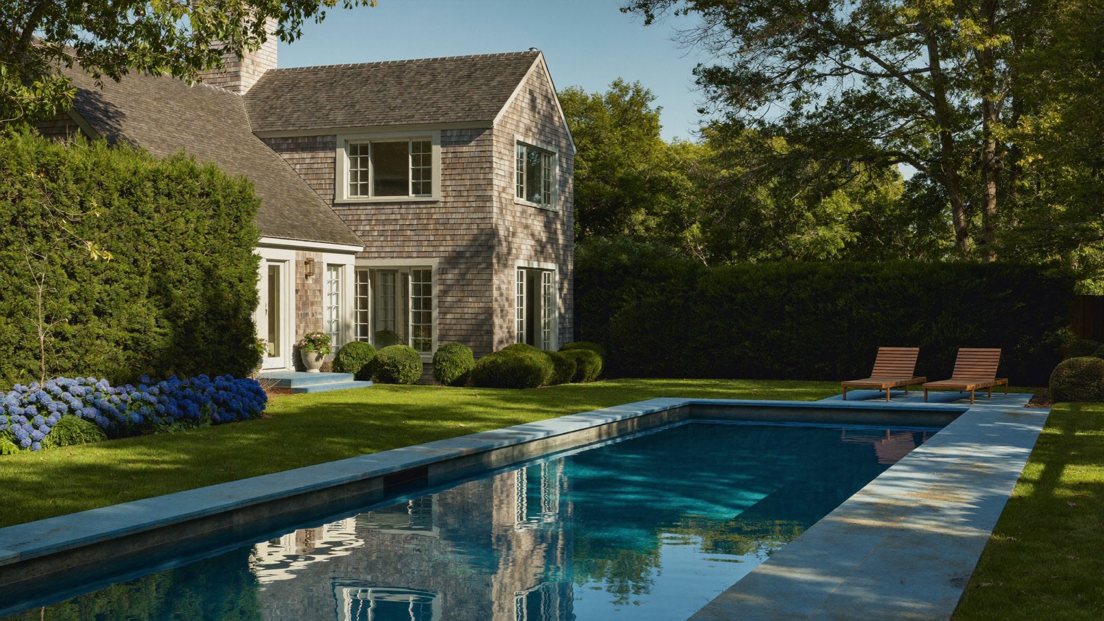 A long pool in late light reflecting a shingled house, framed by a tall hedge, lawn and hydrangeas.