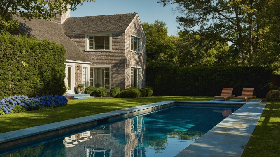 A pool in late light reflecting a shingled house and tall hedge.